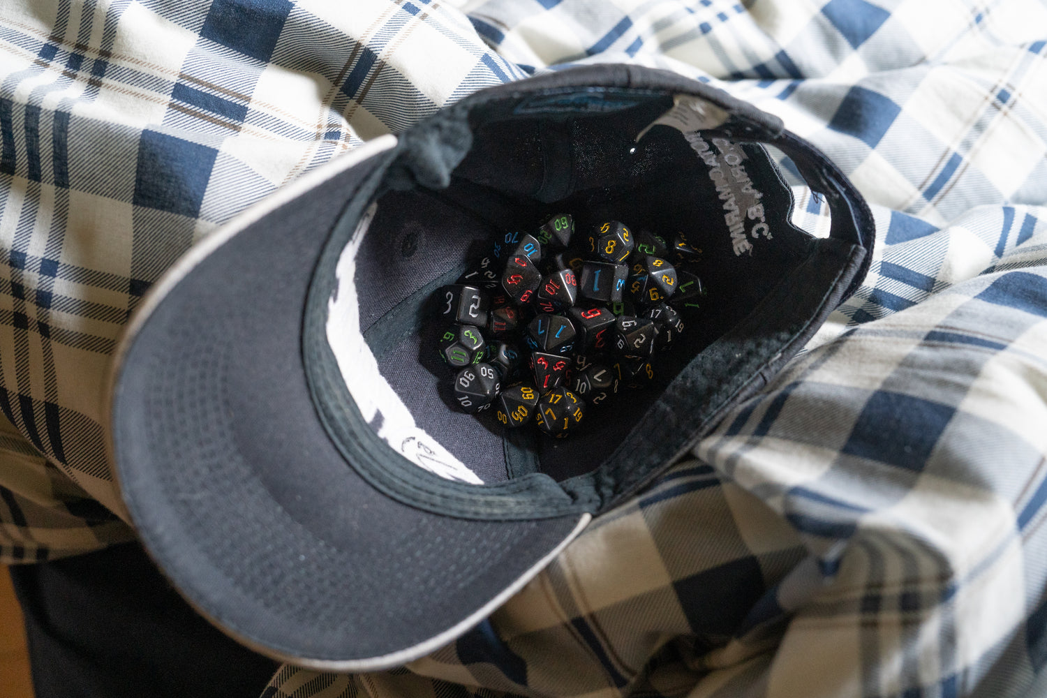 Gray cap containing cursed dungeons and dragons (aka dnd or d&d) game dice sets, including 1d20, d12, d10, d8, d6, d4, all on a plaid fabric bed.